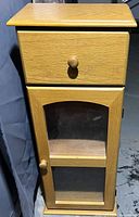 Front view of the wooden storage cabinet showing drawer on top and glass-paneled door with shelf below.