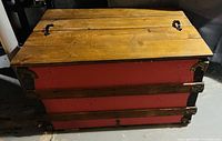 Front top angled view of wooden storage chest showing natural wood plank top, black metal handles, red painted sides, and metal reinforced corners.
