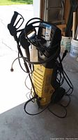 Yellow Karcher electric power washer with black hoses, spray gun, detergent bottle, and power cord, positioned upright on wheels.