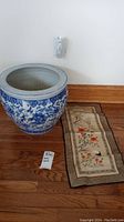 Large blue and white ceramic pot and embroidered fabric art on floor against wall