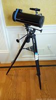 Full view of the Tasco telescope mounted on its tripod stand in a room with wood floor and wallpapered walls.