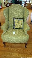 Front view of wingback chair upholstered in light green leafy patterned fabric with needlepoint decorative pillow.