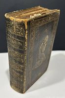 Front angled view of the small, black leather-bound 1849 Holy Bible with gold embossed kneeling praying figure and decorative borders on cover, spine showing 'Holy Bible 1849'