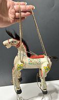 Front and side view of the white wooden marionette horse showing hand-painted floral and geometric patterns, red bridle and saddle, black hooves, real horse hair mane and tail, and attached ropes for control.