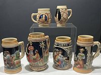 Front and side view of six vintage beer steins on display, showing detailed hand-painted and relief designs with traditional scenery and motifs.
