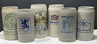 Front view of six vintage German beer steins showing various logos and designs including Albrisbach Klosterbier, Lowenbrau, Black Forest scene, Weihenstephan, Konigsbromer, and Wurzburger.