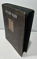 Closed album at slight angle showing textured faux-leather cover and visible gilt lettering “POST CARD ALBUM”