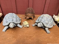 Photo showing three turtle figurines on a wooden surface with a brown wood wall background.