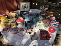Wide shot of 24 collectible tins displayed on a white cloth outdoors. Includes Cracker Jack, Barbie, Monopoly, Coca-Cola, Curious George, and various themed tins.