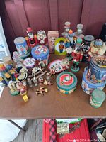 Photo of various collectible metal tins in round, cylindrical and rectangular shapes with vintage artwork placed on a table. Several nutcracker wooden figurines stand upright among the tins. Small collectible figure items and decorative boxes also on table.