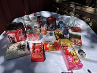 Full overview of the Coca-Cola collection and Barbie dolls laid on a white cloth surface in daylight showing multiple collectible items and two boxed Barbie dolls.