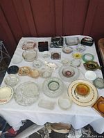 View of assorted ashtrays laid out on white tablecloth outdoors, various shapes, colors, and materials visible.