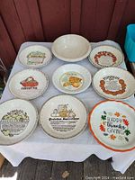 Photo showing eight pie plates on a white cloth on a table outdoors. Plates display various pie recipes including Apple, Cherry, Peach, Pumpkin, Grape, and Quiche Lorraine, plus a Happy Thanksgiving plate. One bowl is plain cream colored.