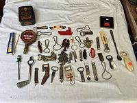 Full assortment of 36 bottle openers arranged on white fabric, showing variety in size, shape and material