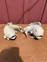 Two fish figurines on a wooden brown table against a red wooden wall background. Fish have open mouths, rounded eyes and detailed fins and scales, made of gray stone or concrete.