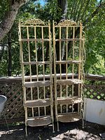 Two tall metal shelving racks with decorative tops and three wicker style shelves each, placed outdoors against a wooden lattice fence with trees in the background.