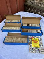 Photo of five blue boxes filled with baseball cards on a white table outside near wooden structure