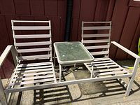 Two white metal lounge chairs with armrests connected by a small rectangular table with a glass top, set outdoors on a wooden deck with brown wall background.