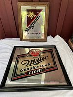 Two vintage bar mirror signs, Magnum Malt Liquor and Miller Genuine Draft Light, placed against a wall on a white cloth, showing the full signs and frames.