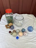 Full collection showing jar with marbles, clear bowl, and various sized decorative spheres on white cloth background against wooden wall.