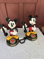 Pair of vintage Mickey Mouse rotary dial phones on white cloth with wood background. Both phones in standing Mickey Mouse figure design in classic colors.