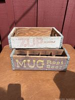 Two vintage beverage crates stacked, one labeled '7 UP' in red on white wood, the other 'MUG Root Beer' in yellow on brown wood with dividers visible.