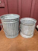 Two galvanized metal trash cans on wooden surface against red wall, shows size and condition.