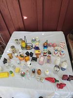 Photo of a collection of assorted salt and pepper shakers arranged on a white cloth surface, including animal, fruit, and novelty shapes in various colors and materials.
