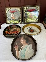 Five vintage signs and trays grouped on a white cloth, two Miller High Life signs, one Budweiser Anheuser Busch tray, one Miller Lite tray, one Coca-Cola tray.