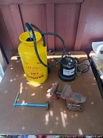 Photo showing yellow Progressive Dura Pack sprayer, black sprayer labeled Lab de Fumigation, two small red bench vises and blue-handled metal tool.