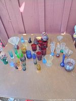 Wide view of the collection showing various colored glass pieces including red, blue, green, amber, and clear barware and vases laid out on a table.