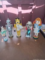 Five Wizard of Oz character dolls displayed standing on a table with a red wooden background.