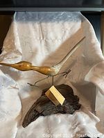 Wooden bird sculpture with long tail mounted on a piece of natural driftwood with a tag attached.