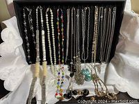 Group of 14 necklaces hanging on black display, showing various styles and materials such as faux pearls, colored beads, chains in gold and silver tones.