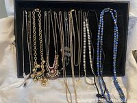 Photo showing 11 various fashion jewelry necklaces arranged on a black tray, including chains, beaded, and rhinestone styles.