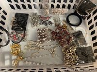 White tray filled with assorted fashion jewelry including necklaces, bracelets, brooches, and earrings, displayed in plastic bags and loose form.