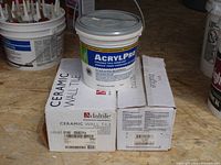 Two boxes of white ceramic wall tile with the top flaps partially open, one tile displayed on top of each box, plus a large white bucket of AcrylPro tile adhesive placed on top of the boxes.