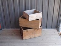 Three vintage wooden fruit crates stacked on a wooden floor against metal siding. Top crate is small and plain light wood. Middle crate is medium size, light brown with faded text. Bottom crate is larger with Blue Bird Canadian Apples branding.