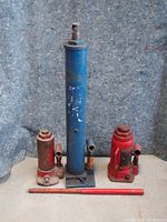 Three bottle jacks displayed side by side on carpeted surface against grey backdrop; largest blue jack with base plate in center, flanked by two smaller red jacks; red two-piece handle laid in front.