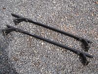 Photo of pair of black universal auto roof racks laid on gravel showing mounting feet and attachment areas
