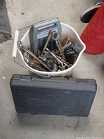 Top view of white bucket filled with chrome sockets, wrenches, and ratchets; gray plastic tool case inside bucket; black plastic tool case on floor