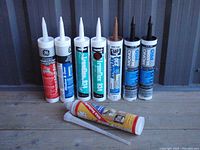 Front view of 7 caulking tubes standing upright on wooden surface.