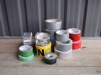 Photo showing assortment of various tape rolls including silver duct tape, green, yellow caution tape, black and white tape on wooden surface.