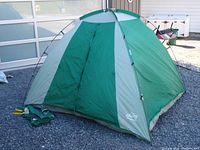 Front view of the green and gray Escape 4-man tent with open zippered door. Stakes and tent pole bag laying beside the tent.