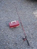 Fishing rod fully extended with red plastic tackle box next to it on gravel surface.