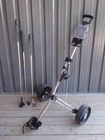 Folding golf push cart with three golf clubs resting beside it on a wood deck.