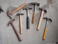 Seven hammers and mallets arranged on concrete floor, showing condition and variety of handles and heads.