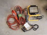 Multiple jobsite work lights including large yellow frame light, smaller floodlight, hanging cage lights and extension cords with light hooks spread on concrete floor.