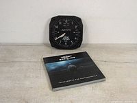Square black Trintec aviation altitude gauge style wall clock with four screws in corners. White altitude markings and numerals 0 to 12 on the dial. Clock hands at 8:20 position visible.