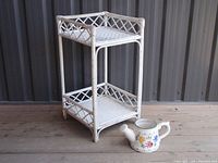 Full view of vintage two-tier rattan plant stand beside ceramic watering can on wooden surface.